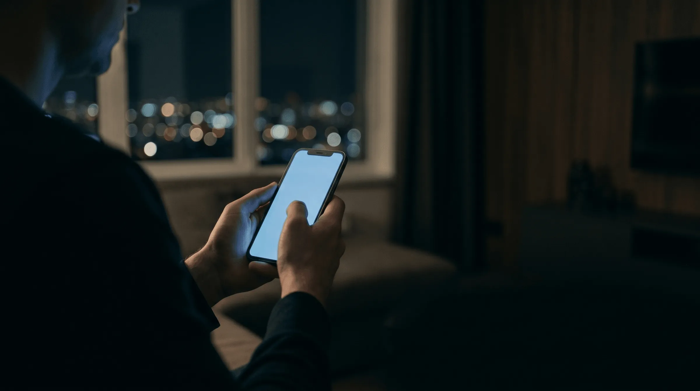 Hands holding a glowing smartphone in an otherwise dark apartment at night, with distant out-of-focus city lights visible through a window behind.